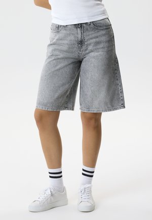 Persoon draagt grijze spijkershorts tot over de knie met subtiele witte spikkels, witte sneakers en witte sokken met zwarte strepen.