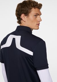 Laivaston värinen polo-paita, jossa on valkoinen chevron-kuvio selkäpuolella. Lyhyet hihat, pehmeä tekstuuri ja perinteinen kaulus.