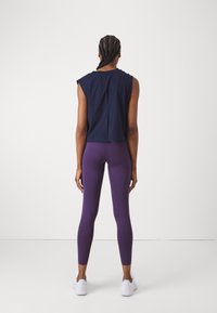 Marineblå kort ermeløs topp med rett nederkant, kombinert med høy-midje lilla tights. Modellen har på seg hvite joggesko.