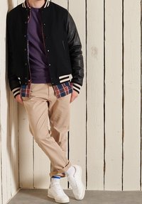 Veste universitaire en cuir noire avec des accents blancs et bleu marine, portée par-dessus une chemise à carreaux. Pantalon beige et baskets blanches complètent le look.