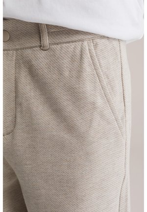 Close-up van beige, gestructureerde broek met een voorzak, riemlussen en metalen knoopsluiting, gecombineerd met een witte ingestopte blouse.