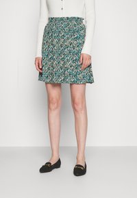 Jupe évasée teal avec un motif beige et vert foncé. Présente une taille élastique. Associée à des mocassins noirs.