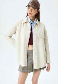 Beige oversized skjortjacka med knappstängning, med en bröstficka, lager på lager över en blå skjorta och en vinröd croptop, kombinerad med en rutiga kjol.