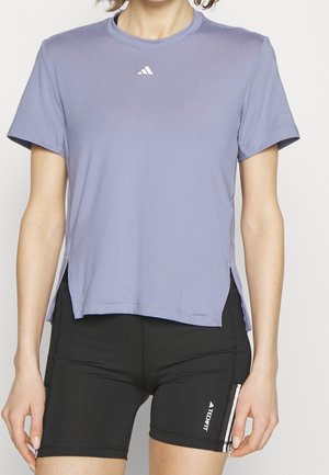 Kobieta ubrana w jasnofioletowy krótki sportowy top i czarne kompresyjne spodenki Adidas Techfit.