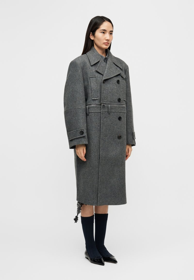 Long manteau en laine grise avec un design croisé à double boutonnage, de gros boutons et une taille ceinturée, associé à des chaussettes bleu marine côtelées et des chaussures noires brillantes.