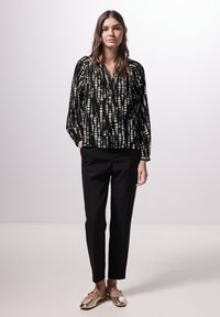 Blusa nera a maniche lunghe con motivo a pois bianchi, abbinata a pantaloni neri su misura e scarpe basse dorate. La blusa presenta un collo a V e una vestibilità rilassata.