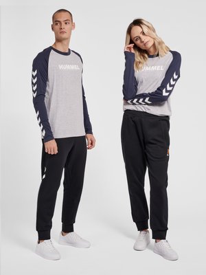 Mand og kvinde står, iført matchende grå og marineblå langærmede skjorter med hvide chevronmønstre og sorte bukser med hvide sneakers.