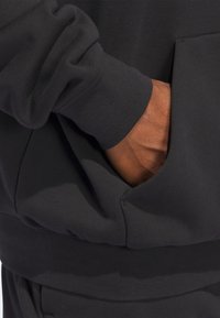 Schwarzer Sweatshirtstoff mit weicher Textur, ausgestattet mit einer großen frontal gelegenen Tasche und gerippten Bündchen, wobei eine Hand in der Tasche ruht.
