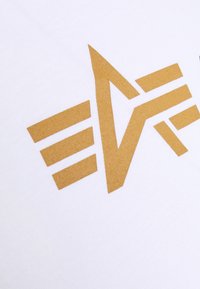 Bílá tkanina se velkým zlatým logem. Logo zahrnuje úhlové linie a pruhy, čímž vytváří odvážný geometrický vzor.