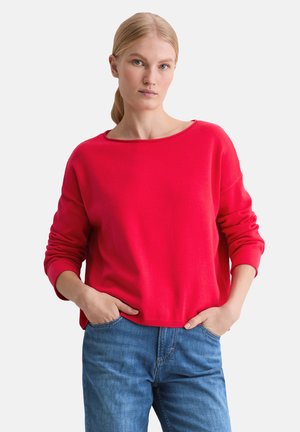 Roter Pullover mit weitem Ausschnitt und langen Ärmeln. Hergestellt aus einem weichen Stoff. Kombiniert mit blauen Jeans, die Hände in den Taschen.