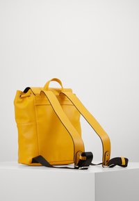 Mochila de cuero amarilla con forma cilíndrica, que cuenta con un asa superior y correas ajustables. Textura lisa con detalles en negro a contraste.