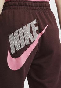 Maroon sweatpants featuring a bold white and pink "Nike" logo and swoosh. Made from soft fabric with an elastic waistband.