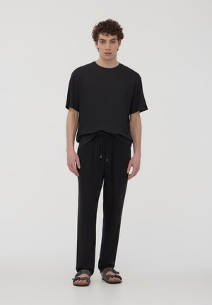 Modello maschile in piedi, rivolto in avanti, che indossa una maglietta nera a maniche corte, pantaloni neri con cordoncino e sandali neri, su uno sfondo chiaro uniforme.