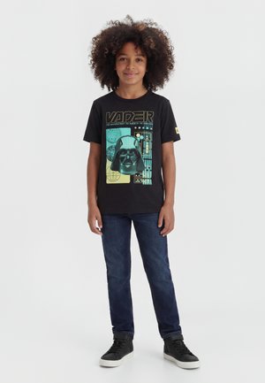 Παιδί με ξανθά σγουρά μαλλιά που φοράει μαύρο μπλουζάκι με τη γραφική παράσταση του Darth Vader, σκούρα μπλε τζιν και μαύρα ψηλά αθλητικά παπούτσια, στέκεται μπροστά σε λευκό φόντο.