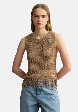 Vrouw draagt een mouwloze, bruin gebreide top met franjes onderaan en lichtblauwe high-waisted jeans, staand tegen een effen achtergrond.