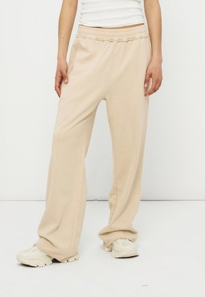 Osoba nosząca beżowe szerokie spodnie z elastycznym pasem i białe trampki, stojąca na jednolitym białym tle.