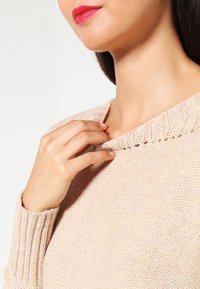 Femme avec du rouge à lèvres rouge ajustant le col d’un pull en maille beige, montrant le décolleté et un détail de la main sur un fond uni.