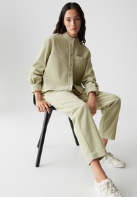Camicia in velluto a coste verde chiaro con colletto alla cinese e taschino frontale, abbinata a pantaloni coordinati. Seduto su una sedia nera, indossa sneakers bianche.