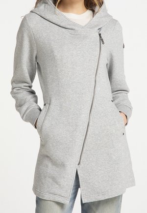 Kvinde iført lysegrå hættejakke med diagonal lynlås og hænder i sidelommerne, kombineret med lyseblå jeans.