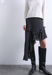 Γυναίκα που φοράει λευκό πουλόβερ, ασύμμετρη μαύρη φούστα με πουά και βολάν, και μαύρες ψηλές μπότες με τακούνι μέχρι το γόνατο, πάνω σε γκρι πάτωμα.