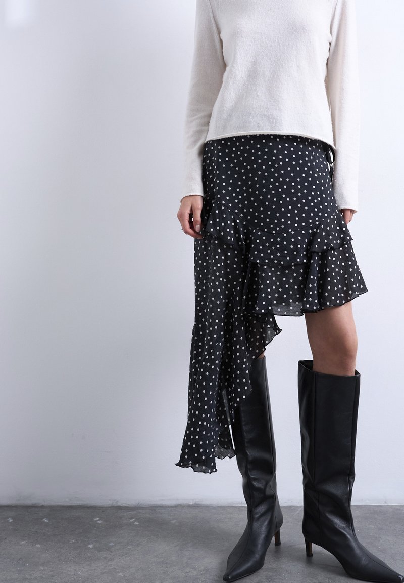 Γυναίκα που φοράει λευκό πουλόβερ, ασύμμετρη μαύρη φούστα με πουά και βολάν, και μαύρες ψηλές μπότες με τακούνι μέχρι το γόνατο, πάνω σε γκρι πάτωμα.