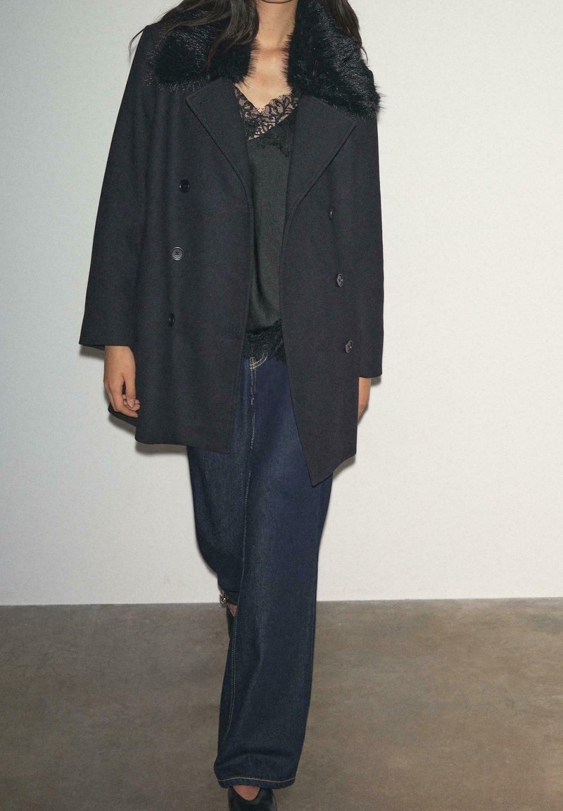 Personne portant un manteau noir croisé avec col en fourrure, un haut noir orné de dentelle, un jean large foncé et des chaussures noires, debout à l'intérieur.