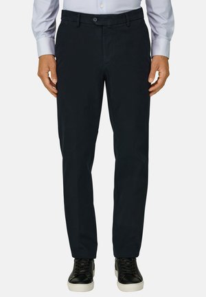 Homme portant un pantalon droit noir, une chemise bleue claire boutonnée et des baskets noires à semelles blanches, debout, bras détendus.