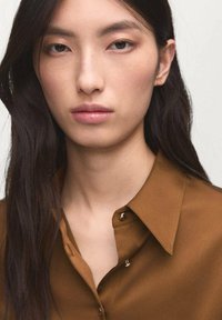 Young woman with long dark hair wearing a brown button-up shirt, looking straight ahead with a neutral expression on a plain background.