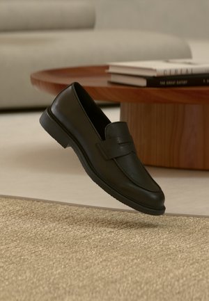 Fekete bőr mokaszin cipő lebeg a levegőben egy bézs szövött szőnyeg fölött, faasztallal és egymásra halmozott könyvekkel egy lágyan megvilágított nappaliban.