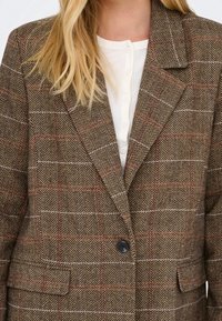 Femme portant un blazer marron à motif chevrons avec un seul bouton noir sur un haut blanc boutonné, avec de longs cheveux blonds visibles.
