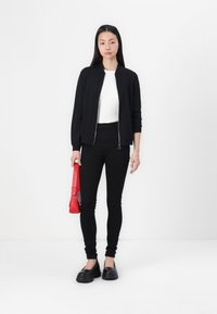 Blouson aviateur noir, haut ajusté blanc, jeans skinny noirs et chaussures noires imposantes. Tenant un sac à main rouge avec une longue bandoulière.