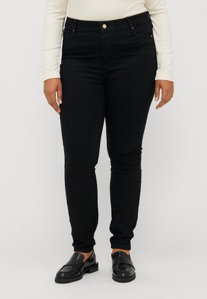 Person trägt schwarze Slim-Fit-Hosen, schwarze Loafers und ein langärmeliges hellbeiges Oberteil, steht vor einem einfarbigen hellen Hintergrund.