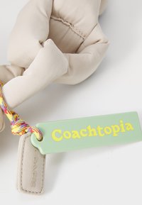 Beige szövet tasak, textúrált felülettel, világos zöld címkével, amelyen a "Coachopia" felirat sárga színben szerepel. Többszínű fonott zsinór jellemzi.