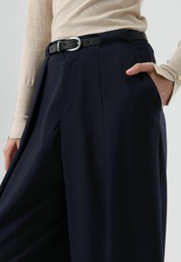 Donkerblauwe broek met wijde pijpen en een zwarte riem met een zilveren gesp, ontworpen met plooien en zijzakken, gemaakt van gladde stof.