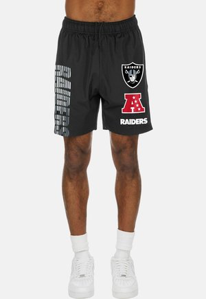 Uomo che indossa pantaloncini sportivi neri con i loghi Raiders e AFC, calze bianche e scarpe da ginnastica bianche, in piedi davanti a uno sfondo semplice.