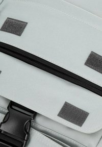 Sac à dos en tissu gris clair en gros plan, avec une fermeture éclair noire, un patch velcro texturé et une boucle noire robuste aux détails de couture visibles.