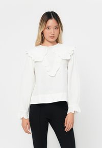Blusa bianca a maniche lunghe con colletto e polsini a volant, realizzata in tessuto leggero, con una linea dritta e dettagli minimi.