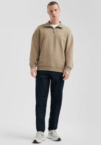 Beige rits sweatshirt met hoge kraag, ontspannen pasvorm, geribde manchetten en een klein logo; gecombineerd met donkere jeans en grijze sneakers.