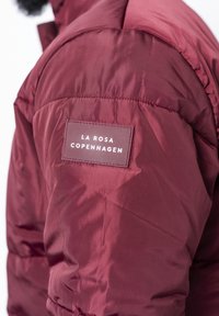 Giacca puffer borgogna con una texture liscia e una patch rettangolare del marchio con la scritta "LA ROSA COPENHAGEN" in bianco. Dettagli della cucitura visibili.