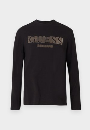 Camiseta de manga larga negra de algodón, con un gran logo metálico dorado "GUESS" en la parte delantera y cuello redondo.