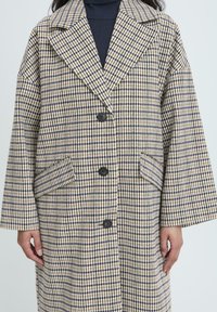 Cappotto in tessuto houndstooth di lana con ampi revers, tre bottoni neri e tasche a soffietto, indossato sopra un maglione a collo alto nero.