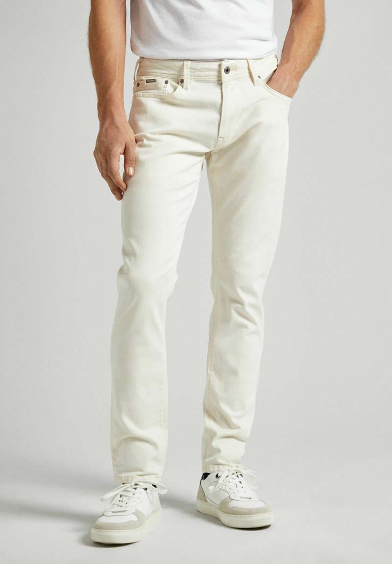 Jean en denim blanc à coupe slim, design à cinq poches, avec fermeture à bouton et à zip standard. Porté avec des baskets blanches, avec des accents gris.