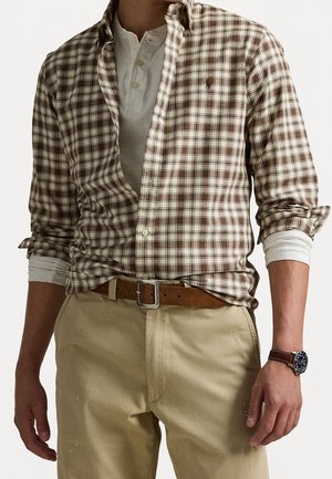 Man met een bruin-wit geruit overhemd met knopen over een wit henley, beige broek, bruine suède riem en een bruine leren horloge met een donkere wijzerplaat.