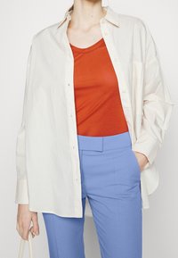 Översized krämfärgad skjorta med knappar över en röd figurnära topp, tillsammans med ljusblå skräddarsydda byxor. Slät textur, enkel design.