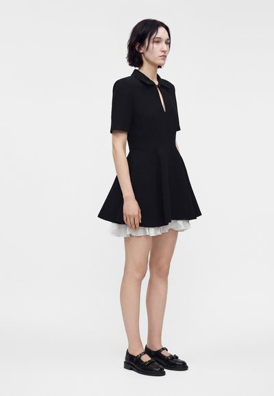 Jeune femme debout portant une robe courte noire avec une jupe évasée, un jupon blanc et des chaussures Mary Jane noires sur fond gris uni.