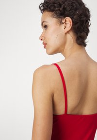 Haut sans manches rouge avec des fines bretelles, présentant une texture lisse et un dos plongeant. De grandes boucles d'oreilles en argent soulignent le look.