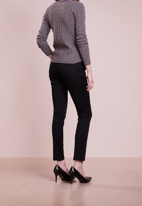 Suéter gris de punto acanalado, pantalones negros ajustados y tacones altos negros brillantes, vistos desde atrás contra un fondo rosa claro.