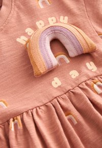 Robe bébé rose douce avec de petits arcs-en-ciel imprimés et une application en tissu arc-en-ciel cousue au-dessus des mots « happy day » sur la poitrine.