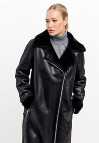 Černý kabát s lesklým povrchem, se sametovým límcem a manžetami, zapínáním na zip a bočními kapsami. Jednoduchý, elegantní design.