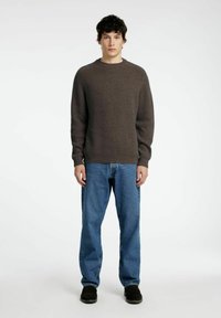 Bruine geribbelde trui met ronde hals, gecombineerd met blauwe spijkerbroeken en zwarte schoenen. Model staat rechtop met het gezicht naar voren tegen een effen achtergrond.